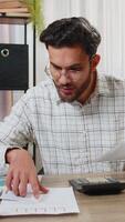 Focused young Indian office worker working with schedules and financial graphics while using calculator sitting at home office desk. Happy businessman freelancer compare and analyze graphs charts. video