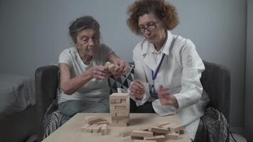 senior kvinna öva Kompetens bygga trä- block, byggnad torn och påfrestande inte till låta den falla, jenga spel. gammal patient dra ut blockera, plats på topp, Stöd läkare under terapi demens i hus. video