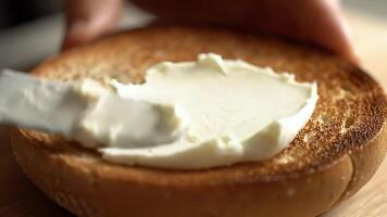 fechar-se tiro do uma pessoa espalhando uma Grosso camada do suave creme queijo para uma dourado torrado bagel metade para uma delicioso café da manhã video