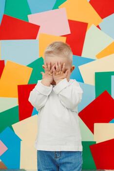 A delightful child is playing, hiding their face behind their hands in a cheerful and playful expression, set against a vibrant and colorful background filled with various shapes and patterns photo