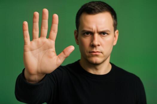 Man Raising Hand in Stop Gesture Against Green Background Indoors Featuring Serious Expression and Closeup Framing With Shallow Depth of Field photo