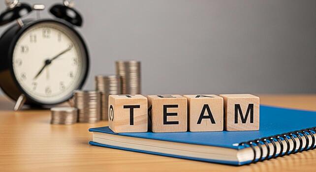 Wooden blocks spelling TEAM placed on a blue notebook with stacks of coins and a clock in the background symbolizing collaboration time management and financial growth in a business setting photo