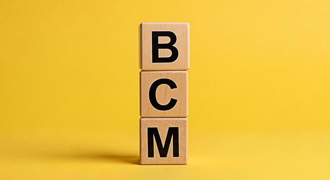 Stacked wooden blocks displaying BCM letters against a vibrant yellow backdrop symbolizing business continuity management and resilience in a simple minimalist studio setting conveying stability and p photo