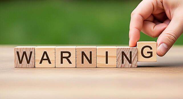 Hand completing the word WARNING with wooden blocks on a table against a blurred green background symbolizing caution and potential risk conveying a message of awareness and safety with a serious tone photo