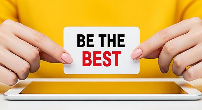 Woman holding a motivational card that says Be The Best in front of a tablet on a yellow background conveying a message of ambition success and selfimprovement in a bright and optimistic setting photo