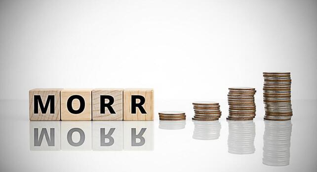 Wooden blocks spelling out MORR Monthly Recurring Revenue next to stacks of coins on a reflective surface representing financial growth and stability in a clean minimalist setting conveying a sense of photo