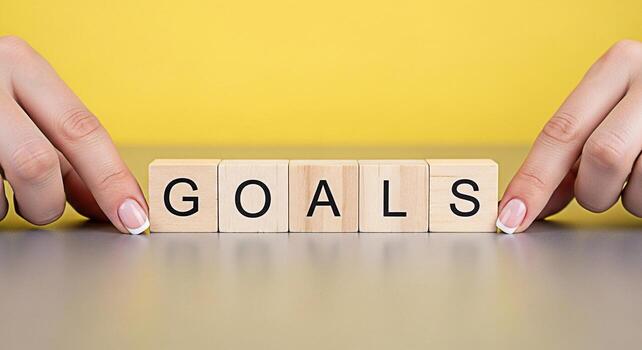Female hands arranging wooden blocks spelling GOALS on a gray table against a yellow background representing ambition achievement and the importance of setting clear objectives for success photo