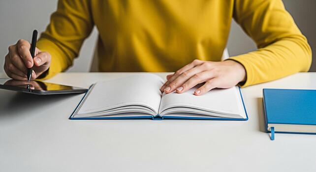 Focused student in a bright yellow sweater writing notes with a stylus on a tablet and in a notebook on a clean white desk conveying a sense of productivity and concentration in a modern learning envi photo