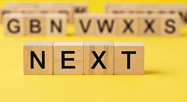Wooden blocks spelling NEXT on a bright yellow surface suggesting anticipation and forward movement with blurred background blocks hinting at a larger alphabet creating a sense of possibility and futu photo