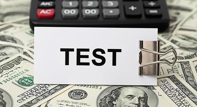 Closeup of a white card with the word TEST displayed held by a silver binder clip resting on a pile of US hundred dollar bills with a calculator in the background conveying a sense of financial assess photo