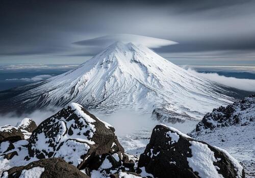 Imposing snow capped volcano summit framed by dark rocks and lenticular cloud photo