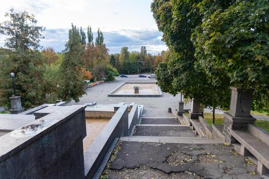 View from the steps of a park with an empty square surrounded by trees and parked cars during a cloudy afternoon photo
