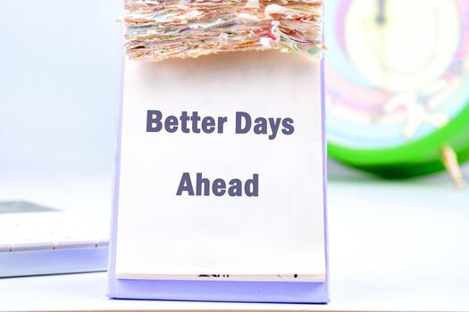Close-up on a calendar displaying the words Better Days Ahead. Stack of used calendar pages are on top, with a colorful clock in the background photo