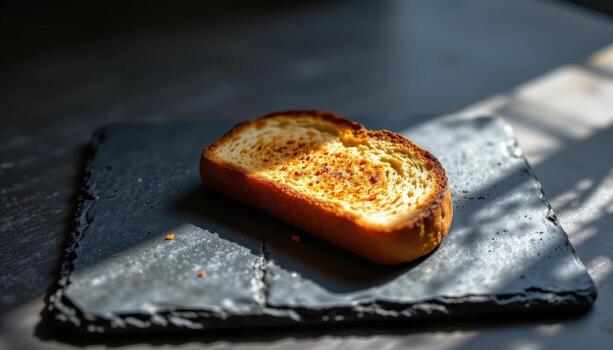 A slice of toast resting on a dark slate surface illuminated by diffused morning light. photo