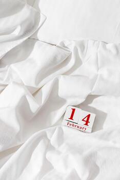 14 february background, holiday date on calendar in crumpled bed with white sheet. Valentine's Day minimal style concept, lifestyle photo. Wooden calendar with red text, vertical, top view, copyspace photo