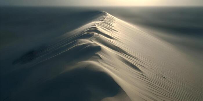 Wind blows across the sharp crest of a sandy desert dune under a hazy sun creating an abstract natural landscape photo