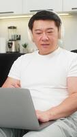 asiatisch Mann beim Zuhause funktioniert auf Laptop mit Ruhe Musik- im Hintergrund bleibt konzentriert und Typen stetig. Chinesisch Kerl auf Sofa Bewertungen Aufgaben setzt Timer Verwendet Umgebungs Klang zum Fokus und lächelt entspannt nach arbeiten. video