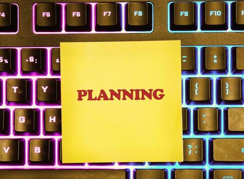 A yellow sticky note with the word PLANNING in red letters rests on a modern keyboard with colorful, backlit keys. It suggests preparation for future endeavors photo