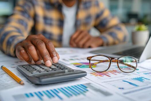 African American man using calculator while analyzing financial documents on desk with laptop and glasses, showcasing focused work in a professional environment photo