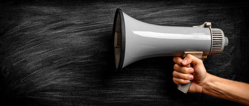Hand holding a gray megaphone against a textured black chalkboard background, symbolizing communication, announcements, and public speaking in a dynamic and engaging manner photo