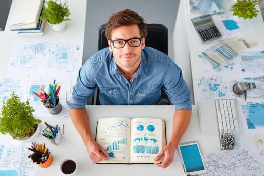 Young man with glasses is analyzing data charts in a notebook while seated at a modern desk surrounded by office supplies and greenery, showcasing a professional workspace photo