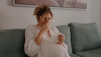 schwanger kaukasisch Frau Niesen und abwischen Nase mit Serviette während Sitzung auf Couch. krank erwartungsvoll Mutter Leiden von Grippe beim heim. video