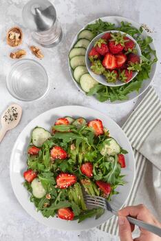A plate of salad stands on a table, top view. A salad of avocado, fresh arugula, cucumber, strawberries, walnuts, and sesame seeds is served on a white plate. A fork takes a bite photo