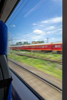 View from train window of red passenger train passing on parallel tracks with motion blur effect under blue cloudy sky, showing railway transportation system photo