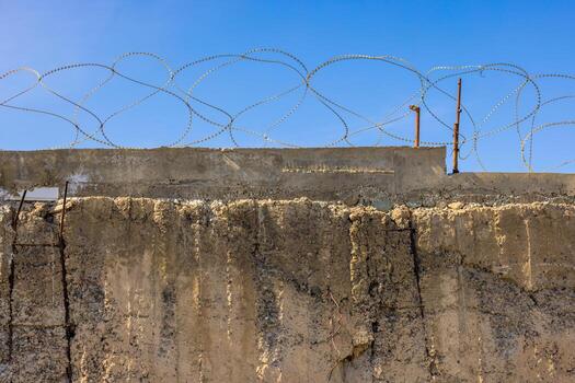 A weathered concrete wall topped with sharp barbed wire under a cloudless blue sky. The rough texture of the wall and the menacing spirals of the wire create a stark and secure boundary. photo