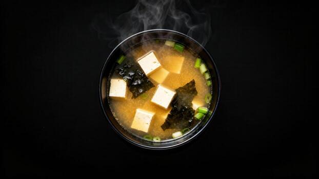 Steaming hot bowl of fermented soybean paste soup with tofu and seaweed floats above a dark surface photo