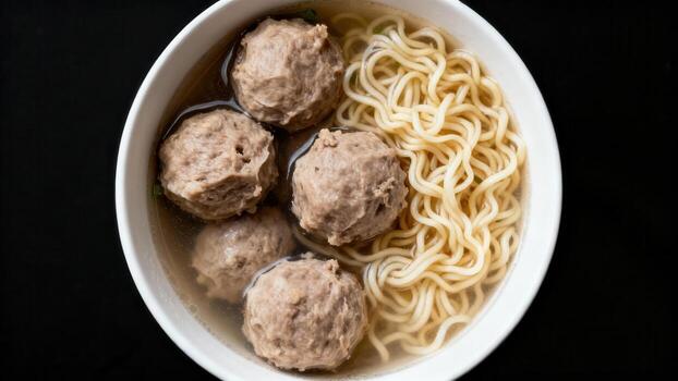 Savory meat spheres and coiled noodles rest submerged in broth within a white ceramic bowl against a dark backdrop photo