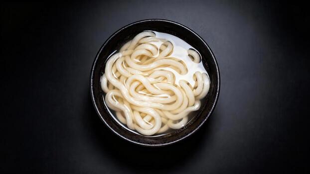 Thick wheat flour noodles submerged in clear broth presented in a dark bowl from an overhead perspective photo