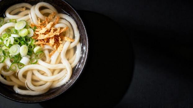 Thick wheat noodles served in a dark bowl with broth, green onions, and crispy fried bits on a dark surface photo