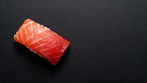 Single piece of fresh fish pressed over rice rests on a dark, textured surface. photo