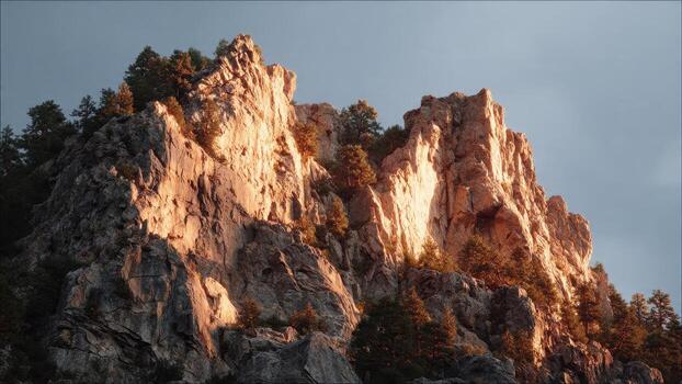 Steep rocky mountain cliff face illuminated by warm sunset light with scattered pine trees under a pale sky photo