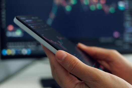 A persons hands cradle a smartphone displaying a stock market analysis chart with red and green indicators, set against a softly blurred backdrop of a larger financial data screen photo
