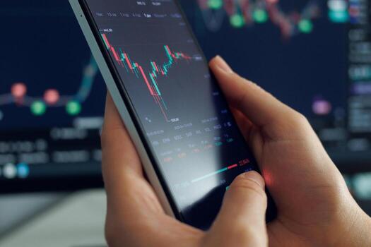 Person interacts with a smartphone showing stock market trends and data analysis, surrounded by additional financial graphics in the background photo