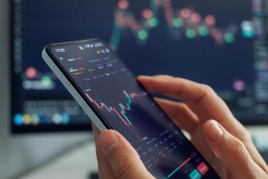 A persons hands hold a smartphone displaying a stock market chart with red and green candles, indicating price fluctuations, against a blurred background showing more financial data on a larger screen photo