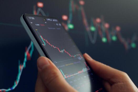 A hand holds a smartphone displaying a declining stock market chart against a blurred background of similar data. photo