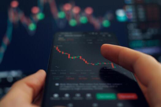 A person interacts with a smartphone displaying stock market data, focusing on financial analysis as graphs update on a nearby computer screen photo