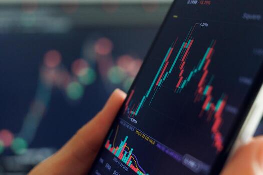 A hand holds a smartphone showing a fluctuating stock chart with red and green candlesticks. Another blurred screen displays similar data in the background. photo