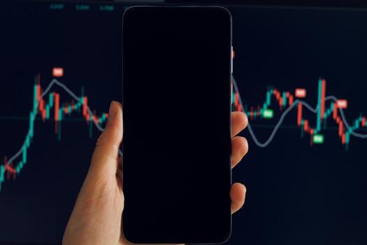 A hand holds a smartphone displaying a fluctuating stock chart with red and green candlesticks. Another blurred screen displays similar data in the background. photo
