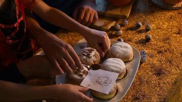 människor framställning för halloween fest förbi dekorera munkar med pulveriserad socker och kakao använder sig av papper schabloner, skapande roligt läskigt ansikten och former på festlig tabell täckt med spindelnät bordsduk video