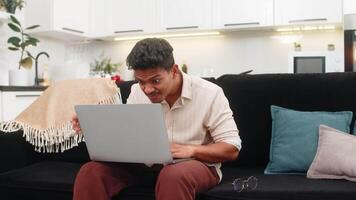 Indian man at home struggles to read tiny text on laptop then puts on glasses and sees clear view. Arabian guy on sofa smiles adjusts frame reads screen lines without strain and continues work task. video