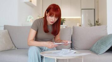 ung kvinna på Hem recensioner papper räkningar och kvitton jämför tal och checkar siffror för fel. rödhårig flicka på soffa innehar penna och kalkylator rynkar pannan på kosta total och verifierar kostnader och datum video