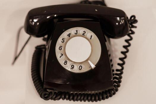 A classic rotary phone with a circular dial and coiled cord rests on a flat, smooth surface. The black exterior adds a nostalgic touch, reminiscent of mid-20th century communication photo