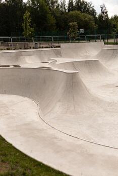A modern skatepark features smooth, flowing concrete structures with various ramps and bowls, ideal for skateboarding and BMX activities. The park is surrounded by green trees and is well-maintained photo