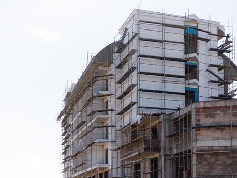 Workers are busy on a multi-story building construction site surrounded by scaffolding. The structure shows progress with newly installed windows and a smooth wall finish photo