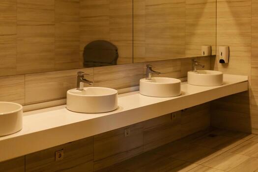 This scene showcases a modern restroom featuring three minimalistic sinks with elegant faucets. The design emphasizes clean lines and a spacious feel, complemented by a large mirror above the sinks photo