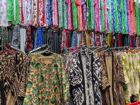 A variety of colorful patterned fabrics and garments are displayed on hangers in a market or store. The textiles feature diverse prints, including geometric, floral, and abstract. photo